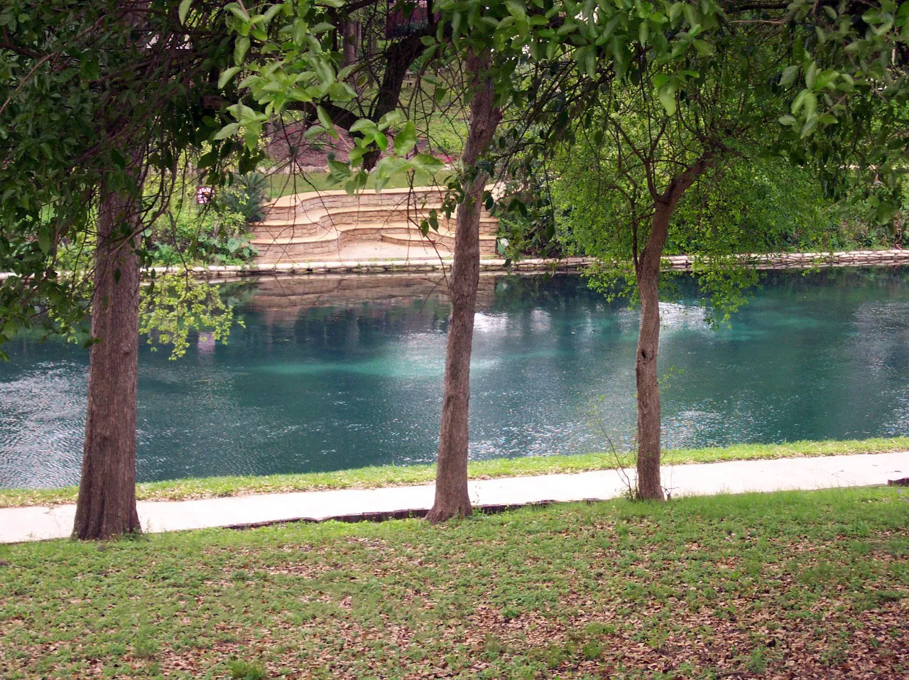 The Comal River in New Braunfels, seen across a lawn between shade trees, with stone steps on the far bank.